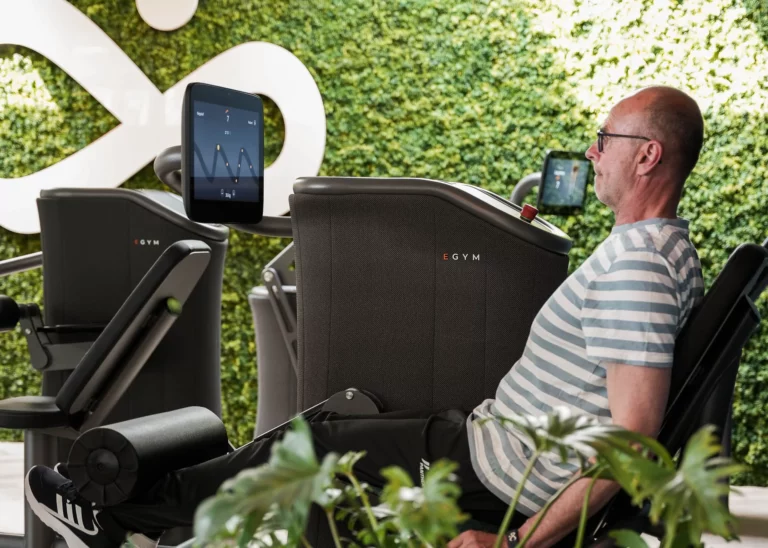 Lid van Sport Connect traint op een EGYM krachttraining toestel. Het logo van Sport Connect is op de achtergrond te zien, evenals planten en groen.